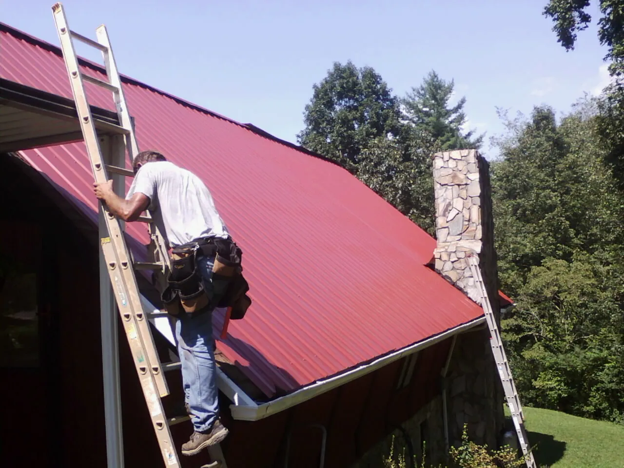 Roofer on a ladder beside a red metal roof and stone chimney — Soffit & Fascia Repair by Roofing Brothers in Ava